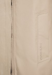 Tela beige con un bolsillo vertical en el lado derecho, bordado con "DIDRIKSONS" en hilo a juego.