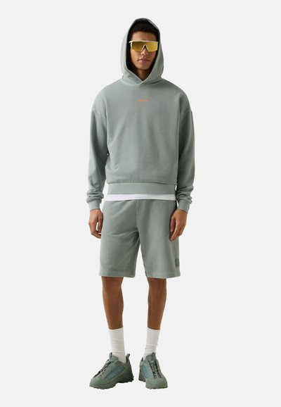 Junger Mann trägt hellgrauen Hoodie mit Kapuze, passende Shorts, weiße Socken, klobige Sneakers und große, gelb getönte Sonnenbrille, stehend.
