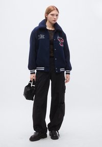 Tamsiai mėlynas koledžo stiliaus švarkas su dirbtinio kailio apykakle, baltais dryžiais ant rankogalių, siuvinėtu logotipu, dėvimas ant juodos raštuotos palaidinės ir kario stiliaus kelnių.