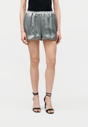 Γυναίκα που φοράει λαμπερά ασημί σατέν σορτς και μαύρα ψηλοτάκουνα σανδάλια με λουράκια στον αστράγαλο, στέκεται μπροστά σε απλό λευκό φόντο.