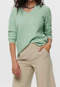 Femme portant un pull en maille côtelée vert clair et un pantalon beige, avec une main dans la poche et un collier chaîne visible.