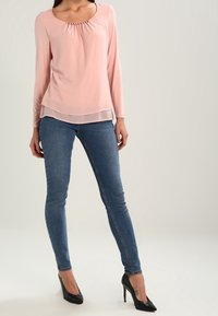 Jasnoróżowa bluzka z długim rękawem, o kwadratowym dekolcie i marszczonym wykończeniu, zestawiona z ciemnoniebieskimi wąskimi dżinsami i czarnymi szpiczastymi szpilkami.