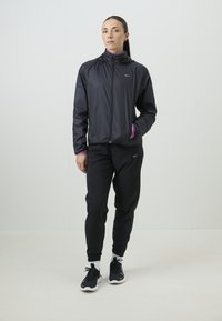 Sort letvægtsjakke med hætte og lilla detaljer, parret med sorte textured bukser og sorte sneakers med hvide såler.