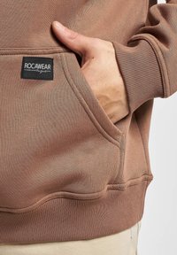 Felpa in cotone marrone con tasca a marsupio, polsini e vita a costine. Presenta un'etichetta nera ROC A WEAR sulla tasca.