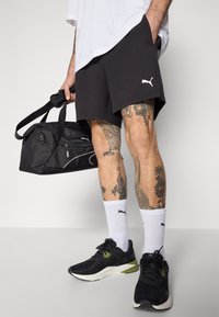 Shorts de sport noirs en coton, arborant un logo Puma blanc. Assortis avec des baskets noires et des chaussettes blanches, portant un sac de sport noir.