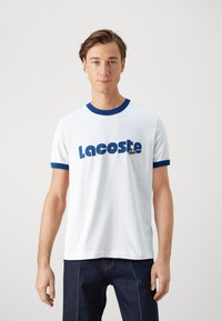 Camiseta de algodón blanca con cuello y pu�ños acanalados azules, con texto en azul en negrita "Lacoste" y un pequeño logo de cocodrilo verde.