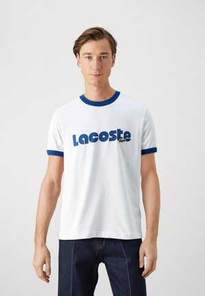 Camiseta de algodón blanca con cuello y puños acanalados azules, con texto en azul en negrita "Lacoste" y un pequeño logo de cocodrilo verde.