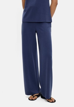 Osoba nosząca granatowe spodnie o szerokich nogawkach, dopasowaną bluzkę i czarne klapki, stojąca na jednolitym białym tle.