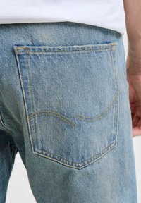 Bolsillo trasero de unos jeans de denim azul claro con costuras naranjas curvas, usados con una camisa blanca.