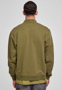 Mand med kort bølget hår iført en olivengrøn sweatshirt over en lysere grøn skjorte, vendende væk mod en ensfarvet grå baggrund.