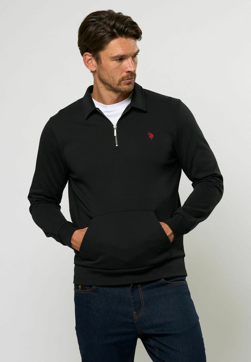 Melna sporta hoodie ar rāvējslēdzēju apkakli, priekšējo kabatu un mazu sarkanu logotipu uz krūtīm. Izgatavots no mīksta materiāla, ar pieguļošu dizainu.