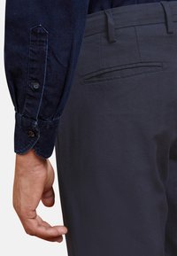 Chemise en denim bleu marine à manches longues avec des poignets à boutons, associée à un pantalon gris foncé doté d'une poche arrière et d'une texture lisse.