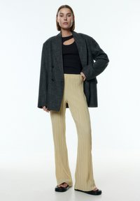 Grijze oversized blazer met dubbele knopen, zwarte aansluitende top en beige geplooide broek met een uitlopende vorm, gedragen met zwarte sandalen.