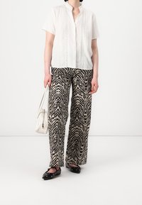 Osoba v bílém krátkém rukávu a kalhotách se zebrovaným vzorem, držící bílou kabelku, s černými plochými botami na jednobarevném pozadí.