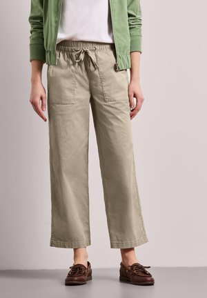 Personne portant un pantalon beige à jambes larges avec des poches avant, une veste verte, une chemise blanche et des chaussures marron à lacets, debout sur un sol gris.