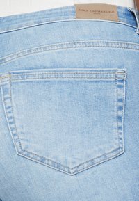 Šviesiai mėlynos džinsinės kelnės su užpakaline kišene, turinčia siūlų apvadu. Diržo srityje matoma šviesiai rudos odos etiketė su prekės ženklo tekstu.