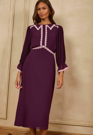 Woman wearing a long purple dress with pleated long sleeves and light pink lace trim on collar, sleeves, and waist standing indoors.