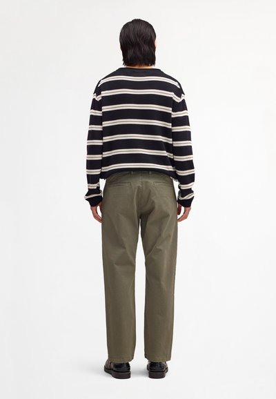 Personne debout de dos, portant un pull à rayures noires et blanches, un pantalon vert olive et des chaussures noires sur fond blanc.
