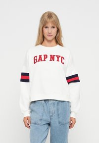 Biała bluza z napisem "GAP NYC" w czerwonym kolorze, granatowe i czerwone paski na rękawach oraz luźny krój. Połączona z jasnoniebieskimi dżinsami.