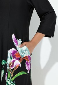 Czarna sukienka z rękawami do połowy przedramienia, ozdobiona żywym kwiatowym nadrukiem w kolorach fioletowym, turkusowym i zielonym. Posiada boczną kieszeń. Gładka faktura.