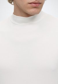 Gros plan d'une personne portant un t-shirt blanc uni à col montant, montrant la partie inférieure du visage et la zone du cou sur un fond blanc.