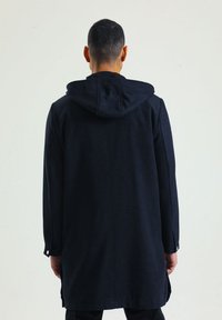 Lange zwarte jas met een capuchon, met knopen aan de mouwen en een rechte snit. Gemaakt van een gestructureerde stof, met een eenvoudig, minimalistisch ontwerp.