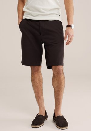 Hombre con pantalones cortos negros hasta la rodilla, camisa blanca texturizada, zapatos marrones sin cordones y un reloj de pulsera, de pie con las manos en los bolsillos.