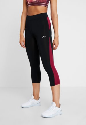 Nainen pukeutuneena mustiin ja tummanpunaisiin lyhennettyihin urheilutrikooihin ja valkoisiin lenkkareihin, seisomassa neutraalia taustaa vasten.