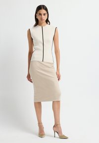 Vrouw in beige midi rok, mouwloos crèmekleurig top met zwarte rand, en beige hoge hakken met enkelbandje tegen een witte achtergrond.