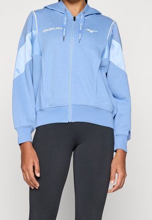 Persona con una sudadera con capucha con cremallera Mizuno azul claro con cordones y leggings negros, de pie frente a un fondo liso.