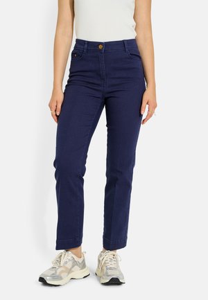 Mujer que lleva pantalones rectos de talle alto de color azul oscuro con botón marrón, top blanco y zapatillas blancas y grises, de pie frente a un fondo liso.
