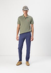 Kaki polo majica s kratkim rukavima i crvenim logom, tamnoplave hlače i svijetlosmeđi tenisice. Sivi šešir upotpunjuje odijelo.