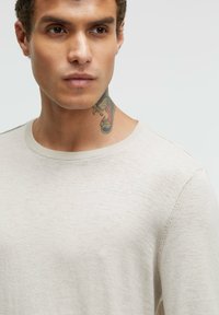 Hellbeige Pullover mit rundem Ausschnitt und strukturiertem Stoff. Eine Nahaufnahme zeigt ein klares Design und sichtbare Nähdetails.