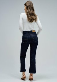 Mujer vestida con jeans oscuros y una camiseta blanca de manga larga, de pie con las manos en las caderas, mirando hacia atrás, sobre un fondo gris liso.