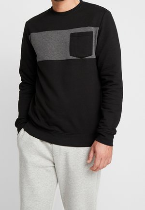 Man wearing a black and gray long-sleeve sweatshirt with a chest pocket and light gray sweatpants, standing with hands relaxed.