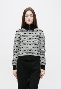 Black and white cropped sweater with zip, featuring striped pattern and "JEANS JOOP!" text. High collar and ribbed cuffs, made from soft material.
