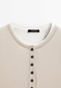 Chemise Henley en coton côtelé beige avec un col blanc, comportant sept boutons noirs sur le devant et une étiquette indiquant "Massimo Dutti."
