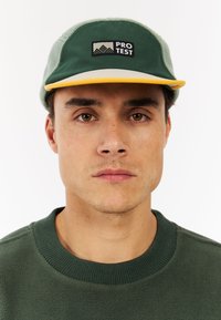 Groene pet met een gele rand, met een zwart "PRO TEST" logo, zachte textuur en een gestructureerd ontwerp. Gemaakt in combinatie met een groene fleecetrui.