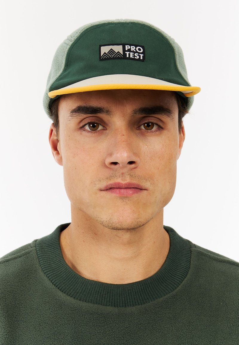 Groene pet met een gele rand, met een zwart "PRO TEST" logo, zachte textuur en een gestructureerd ontwerp. Gemaakt in combinatie met een groene fleecetrui.