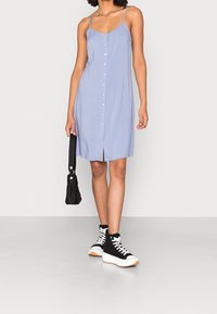 Hellblaues ärmelloses Kleid mit Knöpfen vorne, knielanger Saum, kombiniert mit schwarzen High-Top-Sneakern und einer schwarzen Handtasche. Glatte Stoffstruktur.