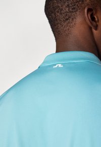 Lys blå polo skjorte med flat krage, med en liten hvit logo på baksiden. Glatt tekstur, tettsittende design.