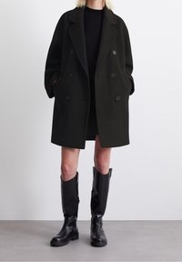 Abrigo de doble botonadura en mezcla de lana de color verde oscuro, con grandes botones, bolsillos laterales y un corte relajado, combinado con botas negras hasta la rodilla.