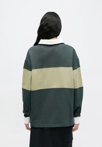 Υπερμεγέθη φούτερ σε σκούρο πράσινο, με οριζόντια χακί ρίγα και λευκό γιακά. Μαλακή υφή, άνετη εφαρμογή και μακριά μανίκια.