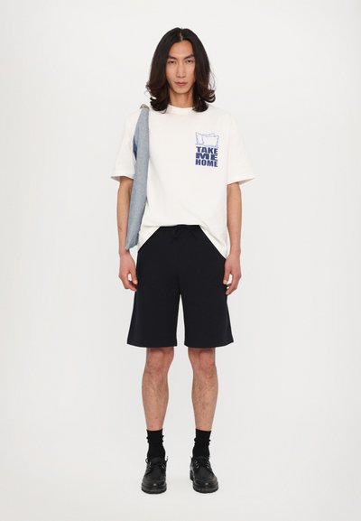 Katoenen T-shirt in gebroken wit met blauwe tekst "NEEM ME MEER," gecombineerd met marineblauwe shorts en zwarte schoenen. Denimjack over de schouder.
