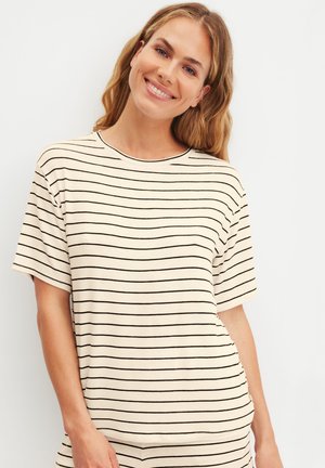 Lächende Frau mit langen blonden Haaren, die ein weißes Kurzarmshirt mit dünnen schwarzen horizontalen Streifen trägt.