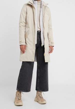 Long manteau d'hiver beige porté sur un gilet matelassé blanc et un col roulé crème, associé à un jean noir à jambes larges et des chaussures de randonnée beiges.