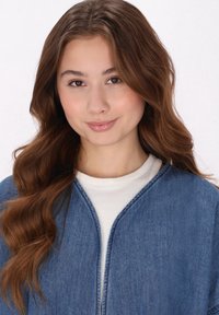 Giubbotto di denim con una tonalità di blu chiaro, dotato di colletto rotondo e apertura frontale, indossato sopra una maglietta bianca a girocollo. Tessuto morbido.