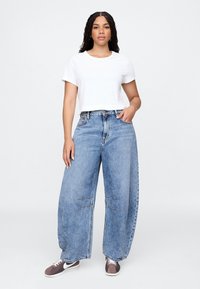 Camiseta blanca corta combinada con vaqueros de pierna ancha en azul claro con detalles cosidos. Lleva zapatillas marrones y blancas.