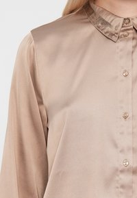 Camicia con bottoni in satin beige chiaro con colletto scintillante in paillettes. Presenta un design classico con una texture liscia e una delicata lucentezza.