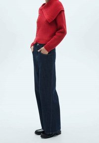 Cardigan en maille côtelée rouge avec un grand col, détails de boutons ; associé à un pantalon en denim large bleu foncé. Chaussures noires avec des accents subtils.